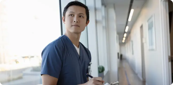 A nurse practitioner in blue scrubs stands by a bright window in a hallway, looking thoughtfully off-camera with an ID badge clipped on.