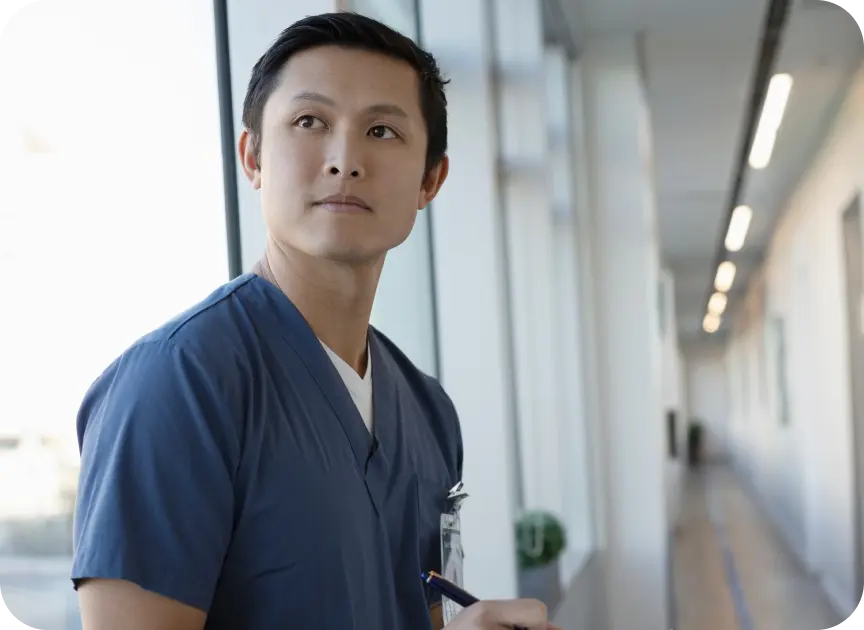 A nurse practitioner in blue scrubs stands by a bright window in a hallway, looking thoughtfully off-camera with an ID badge clipped on.