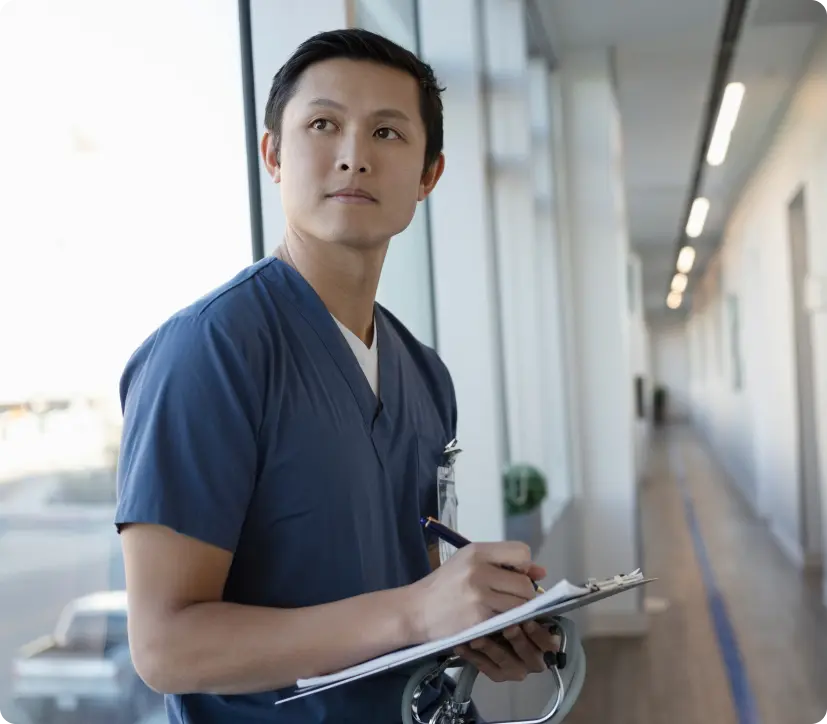 A nurse practitioner in blue scrubs stands by a bright window in a hallway, looking thoughtfully off-camera with an ID badge clipped on.